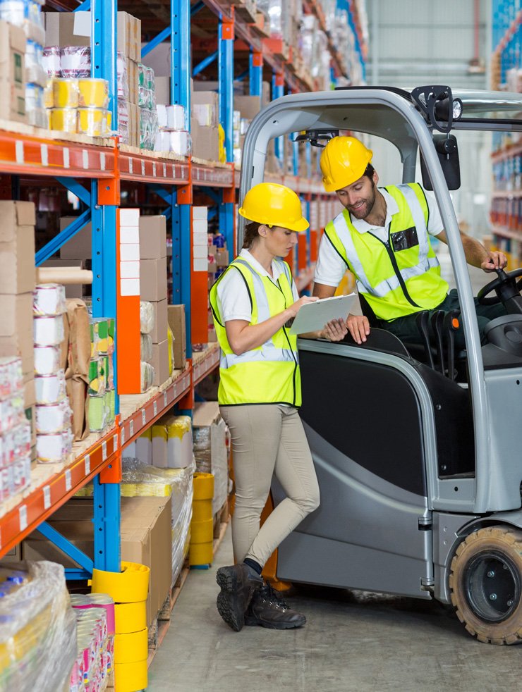 A male forklift operator and a female warehouse worker in safety vests and hard hats discussing inventory while looking at a clipboard in a storage facility.