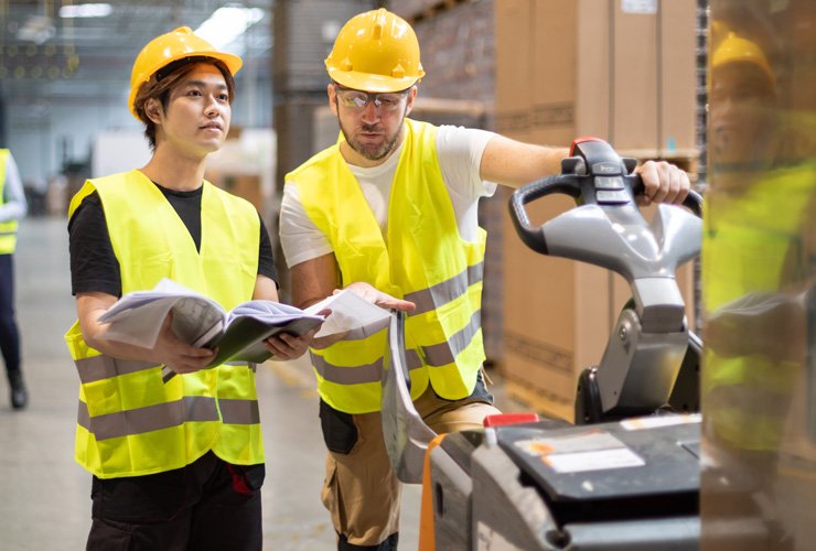 Two warehouse workers wearing yellow safety vests and hard hats reviewing documents while operating a pallet jack in a storage facility with boxes and shelves in the background.