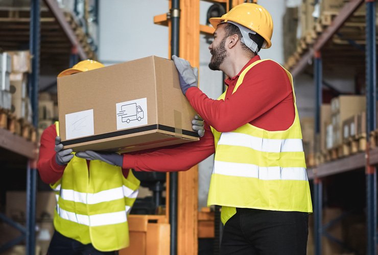 Two warehouse workers in yellow safety vests and hard hats handling a cardboard box with a shipping label, surrounded by shelves filled with inventory in a storage facility.