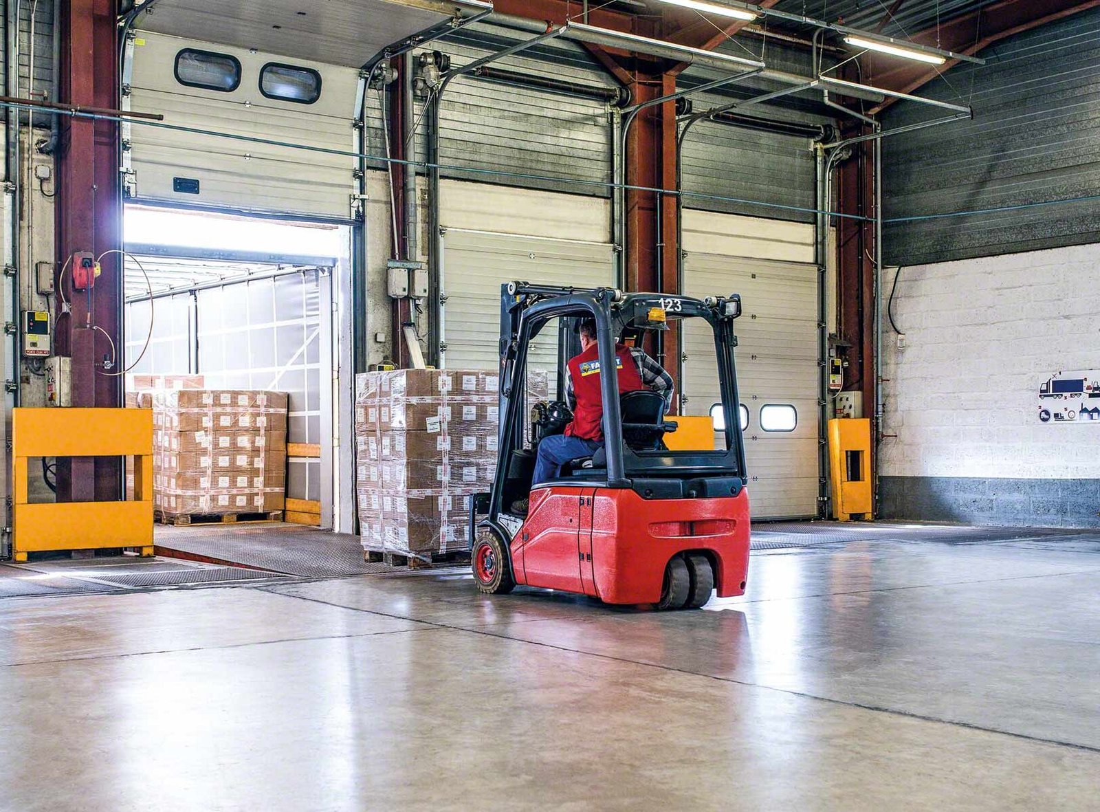 Warehouse worker operating a red forklift to move stacked pallets of goods near an open loading dock door.