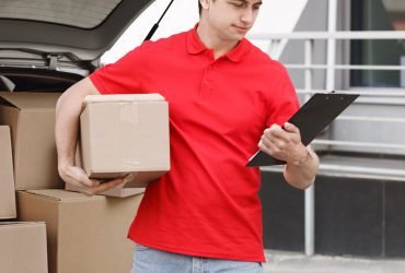 Courier Checking Delivery Details A delivery person in a red uniform holds a package in one hand and a clipboard in the other while verifying shipment details near an open car trunk filled with boxes.