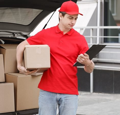 A delivery person in a red uniform holds a package in one hand and a clipboard in the other while verifying shipment details near an open car trunk filled with boxes.